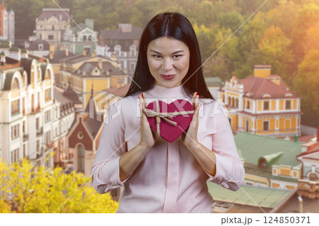 Cheerful asian woman holding a red heart shaped gift box in both hands outdoors. European city background. Cheerful asian woman holding a red heart shaped gift box in both hands outdoors. European city background. 124850371