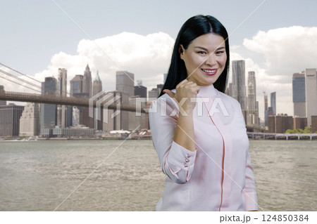 Young asian woman pointing back with her thumb up outdoors. Big city background. Young asian woman pointing back with her thumb up outdoors. Big city background. 124850384