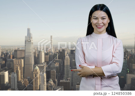 Portrait of a young asian businesswoman with folded arms pointing at copy space. Cityscape with skyscrappers background. 124850386