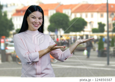 Young cheerful asian woman showing copy space with both hands outdoors. European town scape background. 124850387