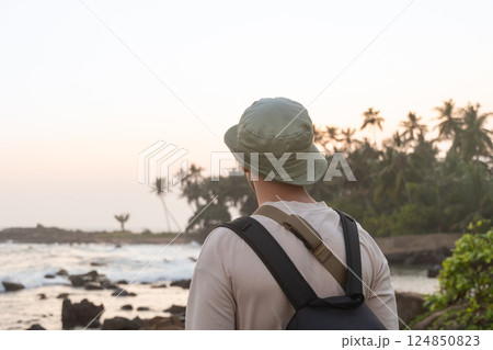 Scenic view of traveler wearing hat and backpack, gazing at ocean during sunset. Palm trees, rocky shore, ocean and serene atmosphere. Travel summer vacation. 124850823