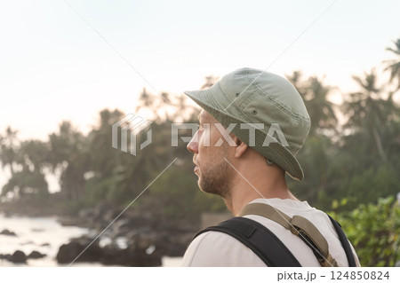 Man in hat and backpack gazing at tranquil tropical coastline during sunrise, looking away. Palm trees and rocks in background. Travel summer vacation. Adventure and reflection. 124850824
