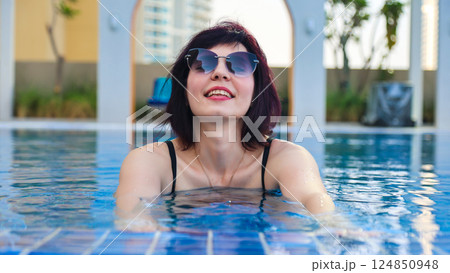 A Woman in a Black Swimsuit Enjoys a Refreshing Swim in a Luxurious Pool, Surrounded by Lush Greenery and Elegant Loungers, Embracing the Serenity and Joy of a Perfect Summer Day 124850948