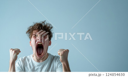 Young caucasian man with messy hair, wearing a white t shirt, screaming with fists raised against a light blue background, conveying intense anger and frustration 124854630