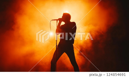 Silhouette of a singer passionately performing live on a stage, illuminated by orange backlighting and surrounded by atmospheric smoke, creating a dramatic and energetic concert scene 124854691