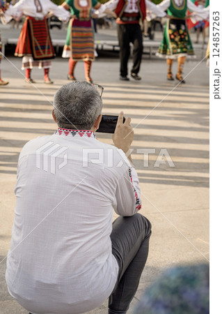 Man squats with smartphone to capture traditional dancers performing in ethnic costumes. People in traditional folk costumes holding hands while dancing in cultural Bulgarian festival 124857263