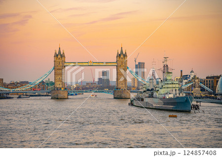 The iconic Tower Bridge stands majestically over the Thames River as HMS Belfast rests nearby. The evening sky glow envelops London, creating a picturesque scene. 124857408