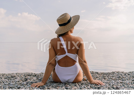 A woman wearing a white bikini and a straw hat is sitting on a beach. The beach is rocky and the sky is cloudy. 124865467