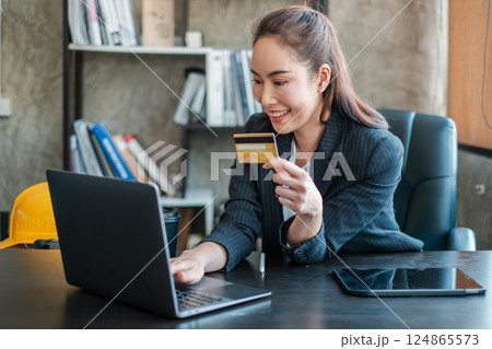 Smiling businesswoman using a laptop and credit card for online shopping in a contemporary office environment. Smiling businesswoman using a laptop and credit card for online shopping in a contemporary office environment. 124865573