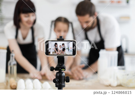 Caucasian family recording cooking tutorial. Parents and daughter knead dough on kitchen table. Caucasian family recording cooking tutorial. Parents and daughter knead dough on kitchen table. 124866815