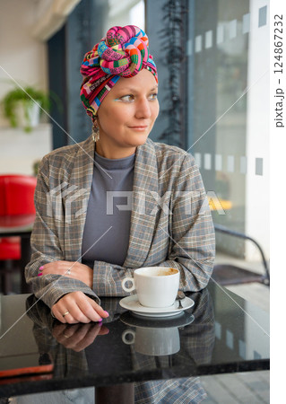 Young woman in a turban sitting in a cafe, drinking coffee and enjoying a peaceful moment. The concept shows that people with illness continue to live, socialize, and express their individuality 124867232