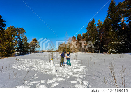 Mature couple constructs snowman together in snowy field near forest, perhaps creating decoration for winter festival or simply enjoying outdoors. Mature couple constructs snowman together in snowy field near forest, perhaps creating decoration for winter festival or simply enjoying outdoors. 124869916