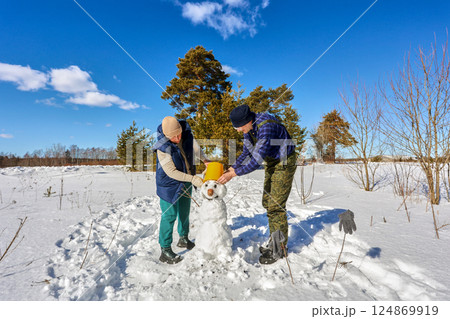 A man and a woman in their 50s are carefully placing a yellow bucket on top of a snowman head in a snowy field surrounded by trees. A man and a woman in their 50s are carefully placing a yellow bucket on top of a snowman head in a snowy field surrounded by trees. 124869919