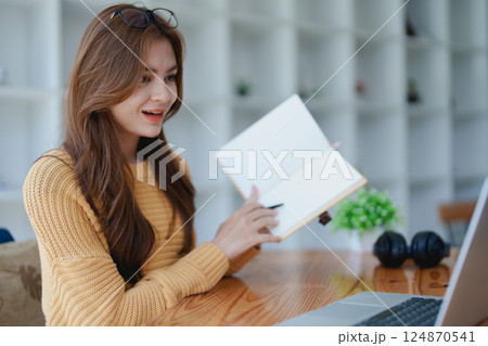 Smiling young woman engaged in a joyful online e-learning video call, gestu while holding a book with headphones on a wooden table nearby Smiling young woman engaged in a joyful online e-learning video call, gestu while holding a book with headphones on a wooden table nearby 124870541