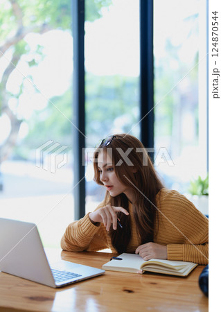Smiling young woman engaged in a joyful online e-learning video call, gestu while holding a book with headphones on a wooden table nearby 124870544