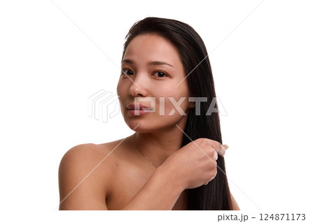 Confident Asian woman in her 20s with flawless skin gently applying product to smooth black hair and combing with brush on white background 124871173