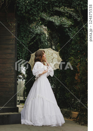 Side view of elegant woman with delicate umbrella under ivy arch, soft glance over shoulder. Suitable for poetic novel or mystery. 124875538