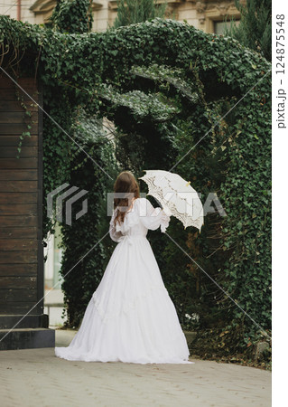 Back view of elegant woman in white Victorian gown holding lace umbrella under archway. Suitable for story or historical fiction. 124875548