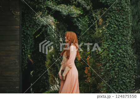 Side view of woman in romantic peach dress standing under green arch, with eyes closed. Suitable for fantasy novel or movie. 124875550