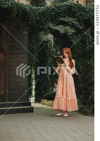 Young woman in peach dress reading antique book near ivy-covered wooden house. Suitable for book or movie cover. 124875552