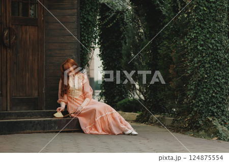 Sitting near wooden door, head bowed, she clasps an open book, vines curling around her like nature's embrace. 124875554