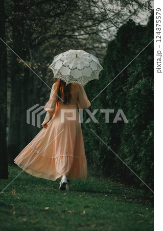 Woman in peach dress walks into fading light under trees with lace umbrella, her silhouette dissolving in dusk. 124875579