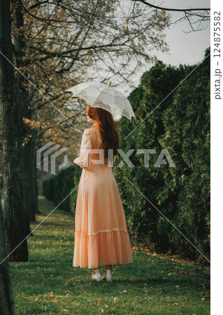 Side view of woman in vintage dress with parasol, caught in afternoon light, pausing in silence with unseen thoughts. 124875582