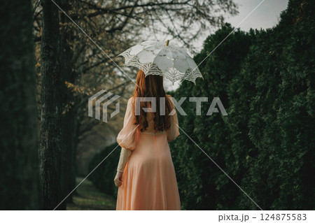 Red-haired woman in peach gown holds lace umbrella, moving between trees and shadows, evoking old-world charm and quiet longing. 124875583