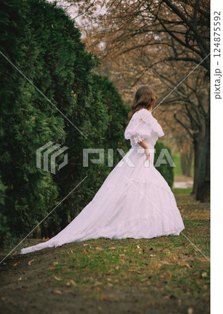 Girl in historical white dress stands still beside green cypress hedge, surrounded by bare trees, lost in thoughts. Suitable for book or film cover 124875592