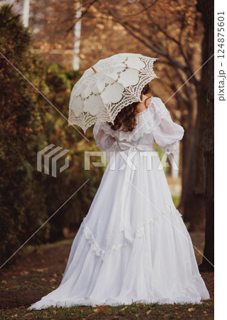 Victorian woman in flowing white gown, looks as princess, standing under lace parasol in golden autumn park. Suitable for film and book covers. 124875601