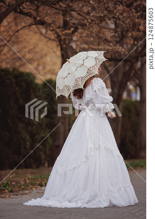 Young woman in white Victorian dress walking away with lace umbrella under autumn trees. Young woman in white Victorian dress walking away with lace umbrella under autumn trees. 124875603