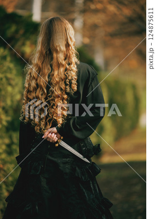 Rear view of woman wearing in black lace dress holding dagger behind back, curly golden hair flowing. Concept of vintage, gothic Rear view of woman wearing in black lace dress holding dagger behind back, curly golden hair flowing. Concept of vintage, gothic 124875611