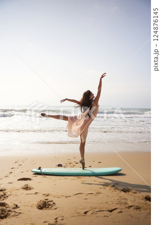 Ballerina in pink dress strikes elegant ballet pose on surfboard by ocean 124876145