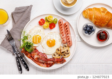 A classic breakfast plate featuring sunny-side-up eggs, crispy bacon, sausages, and fresh tomatoes A classic breakfast plate featuring sunny-side-up eggs, crispy bacon, sausages, and fresh tomatoes 124877023