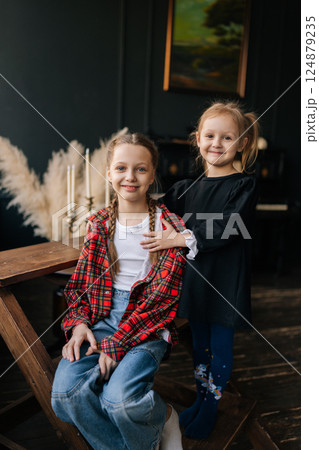 Vertical portrait of vintage-styled sisters sharing close connection, older girl sitting on chair while younger sibling hugging affectionately in warm dark interior setting, looking at camera Vertical portrait of vintage-styled sisters sharing close connection, older girl sitting on chair while younger sibling hugging affectionately in warm dark interior setting, looking at camera 124879235