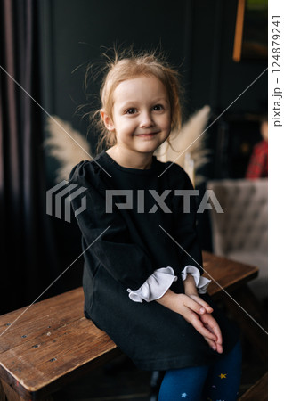 Vertical portrait of pretty young girl wearing ruffled top and blue tights sitting on wooden bench, hands gently clasped, radiating pure childhood innocence with soft smile, looking at camera. Vertical portrait of pretty young girl wearing ruffled top and blue tights sitting on wooden bench, hands gently clasped, radiating pure childhood innocence with soft smile, looking at camera. 124879241