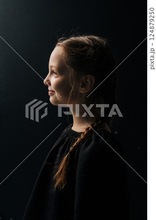 Side view portrait of charming tween girl with braided hair, wearing black dress, posing against dark isolated background in studio. Close-up face of adorable child illuminated by soft light. Side view portrait of charming tween girl with braided hair, wearing black dress, posing against dark isolated background in studio. Close-up face of adorable child illuminated by soft light. 124879250