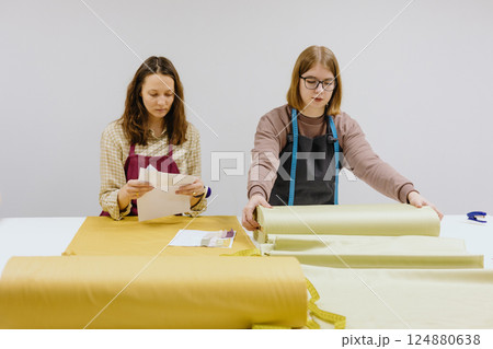 Two female seamstresses and fashion designers stand at table with roll of fabric negotiate Two female seamstresses and fashion designers stand at table with roll of fabric negotiate 124880638