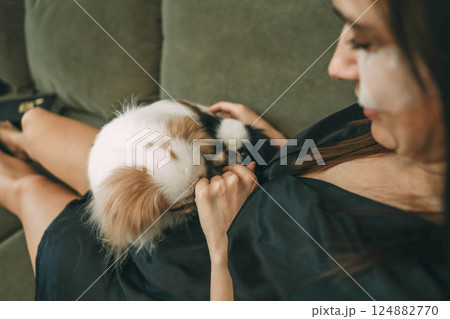 woman in black silk robe relaxing with dogs, smiling while applying under-eye patches, home comfort, concept of skincare and pet bonding 124882770