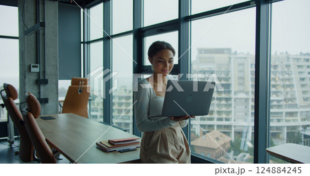 A young woman holding a laptop and working in a modern office with large windows and a panoramic city view, symbolizing professionalism and focus 124884245