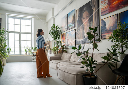 Thoughtful contemplative female artist examines portrait artwork among paintings on studio wall Thoughtful contemplative female artist examines portrait artwork among paintings on studio wall 124885897