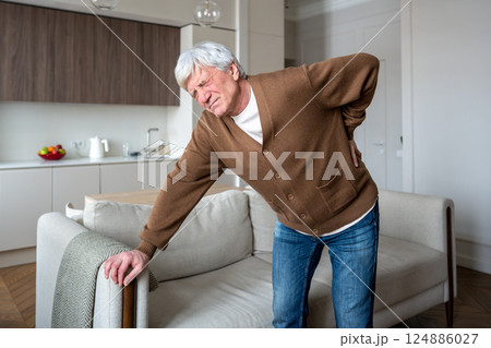 Squinting elderly man suffers alone gripping aching spine with limited mobility in empty house Squinting elderly man suffers alone gripping aching spine with limited mobility in empty house 124886027