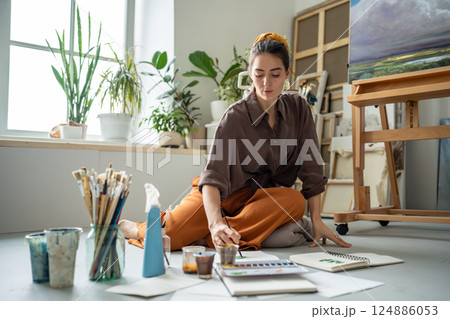 Serious thoughtful female artist paint watercolor in sketchbook on floor in workshop, artwork studio Serious thoughtful female artist paint watercolor in sketchbook on floor in workshop, artwork studio 124886053