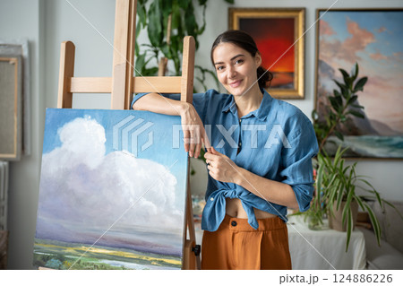 Portrait proud happy woman artist with new painting of sky and nature in workshop, art studio Portrait proud happy woman artist with new painting of sky and nature in workshop, art studio 124886226