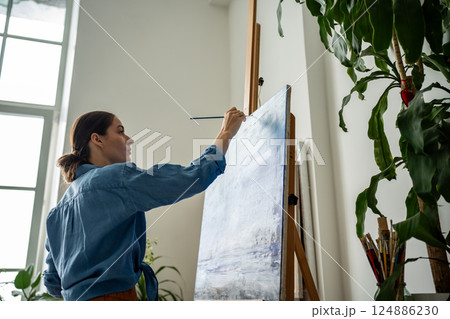 Inspired female artist addding final brushstrokes and signature to landscape painting on easel 124886230