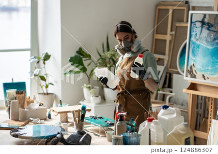 Focused craftswoman in respirator handles reagents preparing epoxy solution for artwork in studio Focused craftswoman in respirator handles reagents preparing epoxy solution for artwork in studio 124886307