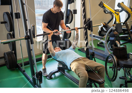 Athletic attentive disabled man in wheelchair works on body strength, lift barbell in gym with coach 124886498