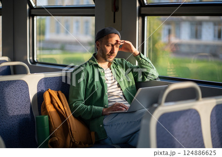 Man works on laptop in train, weary from journey, close eyes to shield from sun. Travel portability Man works on laptop in train, weary from journey, close eyes to shield from sun. Travel portability 124886625