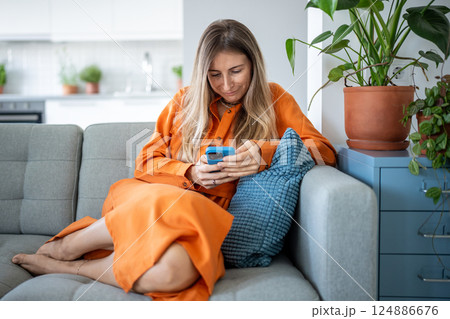 Positive woman resting on couch at home, absorbed in smartphone, enjoying time. Web feed connection 124886676