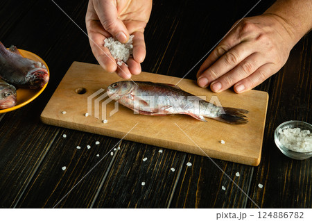 Hands season a freshly caught fish with coarse salt on a wooden cutting board. Nearby, another fish and a bowl of salt add to the kitchen atmosphere, highlighting the pre-cooking process Hands season a freshly caught fish with coarse salt on a wooden cutting board. Nearby, another fish and a bowl of salt add to the kitchen atmosphere, highlighting the pre-cooking process 124886782